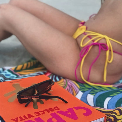 Side view of model sitting on colorful towel wearing Bikini Flavors Strawberry Sunset reversible bermuda bikini bottom.  Yellow reverses to pink.  American made.  Fully adjustable bottoms. 